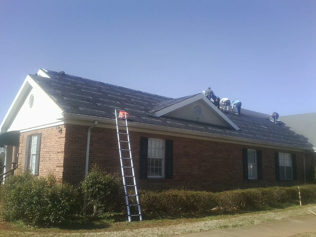 Crew tearing shingles off a brick ranch roof with a ladder set — Metal Roof Repair work by our crew in Weidman