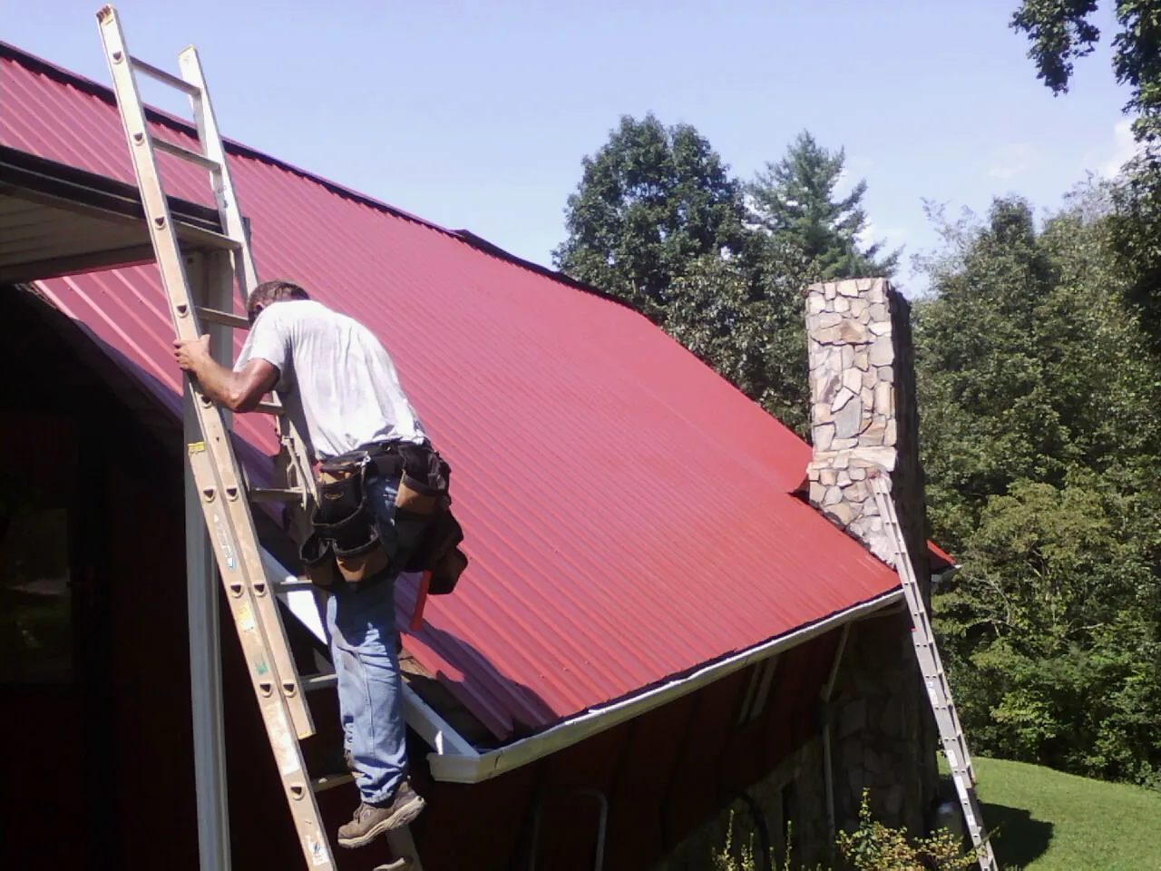 Roofer on a ladder beside a red metal roof and stone chimney — Soffit & Fascia Repair by Roofing Brothers in Weidman